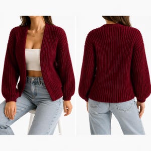 May include: A deep burgundy open-front cardigan featuring a chunky ribbed knit texture, long sleeves with dropped shoulders, and a straight hemline. The garment is shown from both the front and back views, paired with light blue denim jeans.