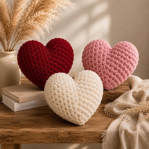 Puede incluir: Tres cojines en forma de corazón hechos de hilo de ganchillo grueso en colores sólidos: uno rojo intenso, uno rosa suave y uno crema. Los cojines están dispuestos juntos, con el corazón crema en el centro delantero, mientras que los corazones rojo y rosa están detrás.