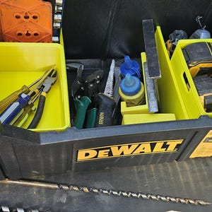 Op de afbeelding: Een zwarte DeWalt gereedschapskist met gele interne opbergbakken en verdelers. De kist bevat diverse handgereedschappen, waaronder tangen, schroevendraaiers en accu's. Een lange metalen boor en een metalen winkelhaak liggen buiten de kist.