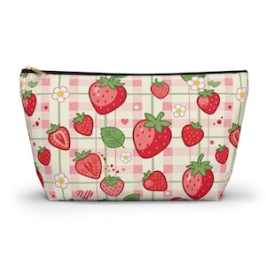 Può includere: Pochette per cosmetici con cerniera con un motivo ripetuto di fragole rosse, foglie verdi e fiori bianchi su uno sfondo a scacchi rosa e bianco. La pochette ha una cerniera nera e un tirante dorato.