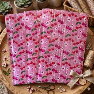 May include: Pink bubble mailers with a repeating pattern of red cherries, green leaves, and white flowers. The mailers are arranged on a wooden tray with decorative elements, including a bow with an "Etsy" tag.