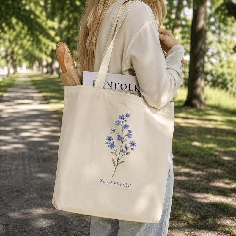 Woman carrying a cream cotton tote bag with blue forget me not flowers and “Forget Me Not” text, inspired by Outlander and Scottish wildflowers, walking through a sunny park.