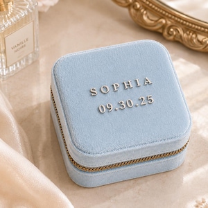 Peut inclure: Une boîte à bijoux carrée en velours bleu clair dotée d'une fermeture éclair dorée. Le dessus de la boîte est personnalisé avec le nom SOPHIA et la date 09.30.25 en lettres métalliques argentées en relief centrées sur la surface en tissu.