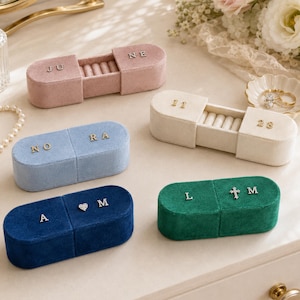 Puede incluir: Cinco joyeros ovalados de terciopelo en rosa, crema, azul claro, azul marino y verde oscuro. Cada caja tiene una tapa deslizante con letras o símbolos metálicos, incluyendo los textos JUNE, 11 28, NORA, A M y L M con una cruz.