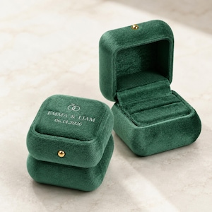 May include: Two dark green velvet ring boxes with a gold-tone metal clasp. One box is open, revealing a matching velvet interior with a slot for rings. The closed box features white text reading EMMA & LIAM 06.14.2026 and a line art ring icon.