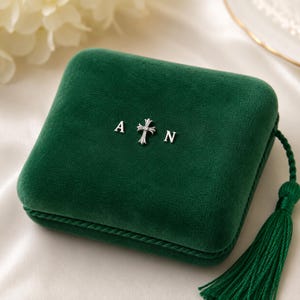 May include: A square, dark green velvet jewelry box featuring a silver-toned metal cross emblem centered between the letters A and N. The box is finished with a matching green braided cord trim and a decorative green tassel attached to the side.
