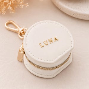 May include: A white, heart-shaped pouch with the name "LUNA" in gold lettering. The pouch has a gold zipper and a gold clip for attaching to a bag or keychain. The pouch is likely for carrying small items.
