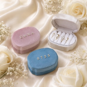 May include: Three velvet ring boxes in pink, blue, and white, with the word "EMMA" and the numbers "0725" in silver lettering. One box is open, displaying rings. The boxes are on a cream-colored satin surface, with white roses and baby's breath.
