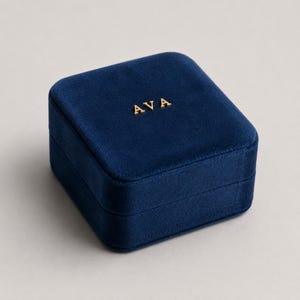 May include: A navy blue velvet jewellery box with the gold letters "AVA" on the lid. The box is square and has a soft, plush texture. The box is closed and sits on a light-coloured surface.