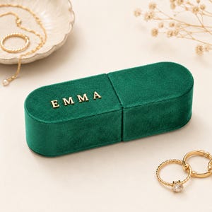 May include: A green velvet jewelry box with the name "EMMA" in gold letters. The box is open, revealing its interior. Gold jewelry, including a necklace and rings, is arranged around the box. The background is a neutral color.
