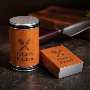 May include: A cylindrical wooden knife sharpener and a rectangular wooden knife sharpener, both featuring a silver top and the engraved text "Ethan's Kitchen" alongside a graphic of a crossed spatula and fork on the light brown wood surface.