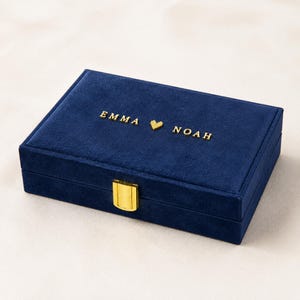 Può includere: Un portagioie rettangolare in velluto blu navy con una chiusura in metallo dorato sul davanti. Il coperchio è personalizzato con il testo dorato EMMA, un piccolo simbolo a forma di cuore e il nome NOAH centrati sulla superficie superiore.