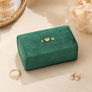 May include: A rectangular dark green velvet jewelry box featuring gold-colored metal letters A and M with a small heart symbol in the center. A gold-toned ring with a clear stone and a pair of matching gold-toned stud earrings are placed nearby.
