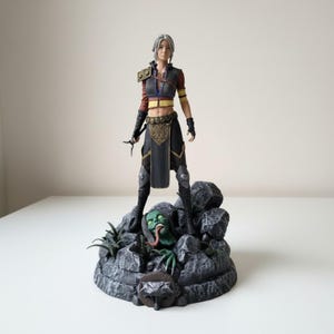 May include: A detailed figurine of a silver-haired warrior standing on a rocky base. The figure wears a dark vest, black trousers, and armor pieces. A green, long-tongued creature head emerges from the rocks at the base, which features a wolf emblem.