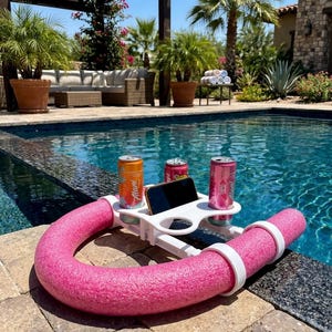 May include: A white plastic floating pool caddy featuring a curved pink foam noodle base, two circular cup holders containing orange and pink aluminum beverage cans, and a central rectangular slot holding a black smartphone device.