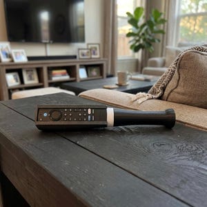 May include: A black remote control holder shaped like a baseball bat. The holder features a textured dark gray casing that encases the remote, revealing the buttons, and transitions into a smooth, solid black handle with a white accent ring.