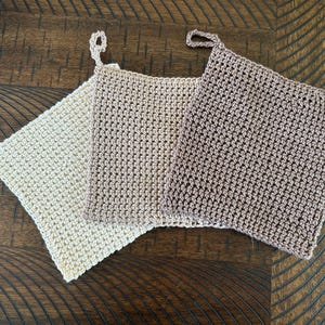 May include: Three handmade crochet potholders in cream, tan, and brown. Each square potholder has a loop for hanging. The potholders are made of a textured yarn and are displayed on a dark wood surface.