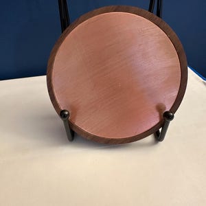 May include: A round coaster with a copper-colored center and a dark wood border. The coaster is displayed on a black metal stand, showcasing its design. The coaster is a decorative item.