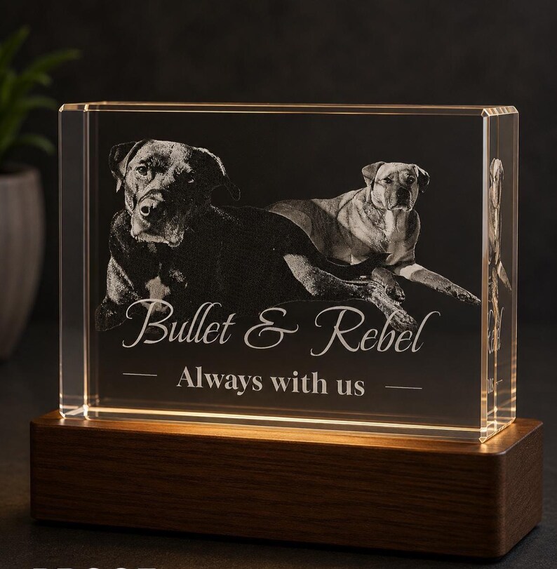 May include: A rectangular clear glass block featuring a laser-etched image of two dogs lying down. The text "Bullet & Rebel" and "Always with us" is engraved below the dogs. The glass block rests on a rectangular dark brown wooden base.