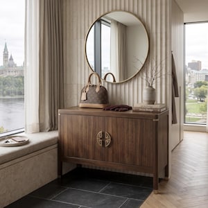 May include: A brown and tan handbag with a luggage tag sits atop a dark wood cabinet. A round gold mirror hangs above the cabinet. A window with a view of a city and a tray with keys are also visible.