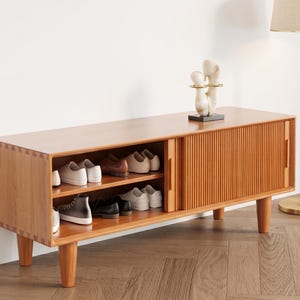 May include: A wooden shoe storage cabinet with a light brown finish. The cabinet has two open shelves on the left side, displaying various pairs of shoes. The right side features a sliding door with vertical grooves. The cabinet is supported by four rounded legs.