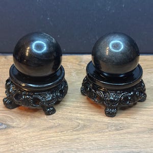 May include: Two smooth, polished black obsidian spheres, each resting on a decorative black resin stand. The stands feature intricate, carved detailing and three small feet. The spheres have a reflective surface with subtle golden sheen highlights.