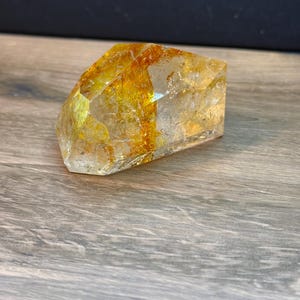 May include: A faceted, clear quartz crystal specimen featuring prominent golden-yellow rutile inclusions. The stone has a polished, angular shape with visible internal fractures and a vibrant, warm-toned mineral vein running through the center.