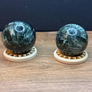May include: Two polished dark green spheres with fibrous, chatoyant white inclusions, each resting on a circular, light-colored wooden stand featuring a sunburst-like carved pattern around the perimeter of the base.