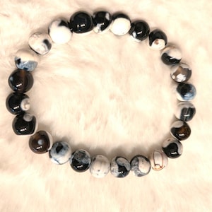 May include: A beaded bracelet featuring round, polished beads in a mix of black, white, and gray. The beads are strung together, forming a circular shape. The bracelet is displayed on a soft, white, textured surface.