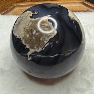 May include: A polished, spherical decorative object resembling a globe. The globe features a dark, glossy surface representing the oceans, with tan and cream-colored continents. The object is resting on a white, textured surface.