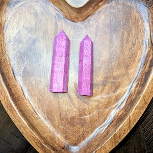 May include: Two pink crystal obelisks rest in a heart-shaped wooden bowl. The obelisks are rectangular with a pointed top. The bowl is made of a light brown wood with a carved heart shape.