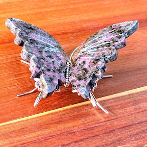 May include: A stone butterfly sculpture with wings in shades of purple, black, and gray, resting on a wooden surface. The butterfly has a silver wire body and legs, and the wings have a textured appearance.