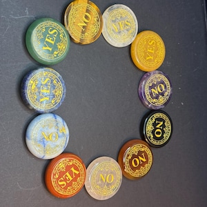 May include: A collection of eleven circular polished stones in various colors, including green, brown, clear, yellow, blue, purple, black, and red. Each stone features a gold-colored border and the word YES or NO printed in gold lettering.