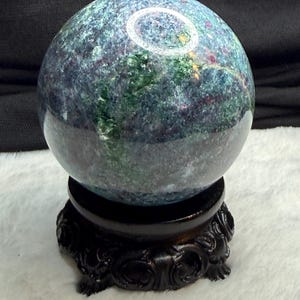 May include: A polished, spherical gemstone with a mottled pattern of blue, green, and red hues. The sphere sits on a dark, ornate wooden stand. The stand has a detailed, carved design.