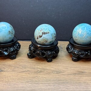 May include: Three polished light blue Larimar spheres with white and brown mineral inclusions, each resting on a black circular display stand featuring ornate, carved detailing and three small feet for stability and support.
