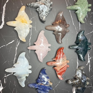 May include: A collection of ten carved stone figurines shaped like manta rays, featuring various colors and patterns including white, grey, green, pink, red, blue, and dark mottled tones, each with a smooth, polished surface and distinct fins.