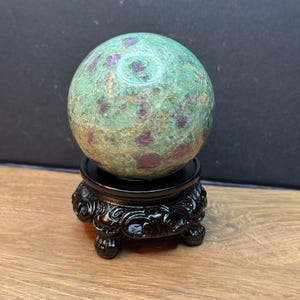 May include: A polished spherical stone featuring a mottled green base with embedded deep magenta and dark red mineral inclusions. The sphere rests securely on a black, circular, carved wooden display stand with three ornate, curved feet.