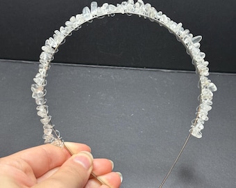 May include: A thin metal headband adorned with numerous small, clear, irregular-shaped crystal-like stones wrapped along the top arch. The headband features two clear plastic protective tips at the ends of the wire frame for comfortable wear.