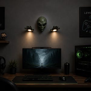 Könnte beinhalten: Ein Gaming-Setup mit einem Computermonitor, einer Tastatur und einer Maus auf einem Holzschreibtisch. Die Wand zeigt einen Alienkopf, handförmige Lichter und Gaming-Kunst. Ein Gaming-PC, Kopfhörer und eine kleine Pflanze sind ebenfalls zu sehen.
