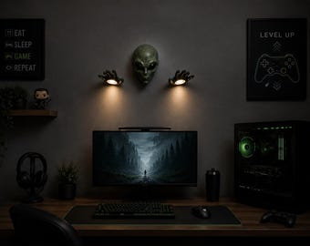 May include: A gaming setup featuring a computer monitor displaying a forest scene, a keyboard, mouse, and a gaming PC. The wall is decorated with an alien head, hand-shaped lights, and framed gaming-themed art. A shelf holds a figurine and a plant.