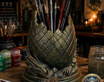 May include: A detailed, green dragon egg-shaped pen holder with a textured surface, holding various art brushes and pens. The base features a dragon design and a bronze-colored emblem. The holder is on a wooden surface, with paint supplies visible in the background.