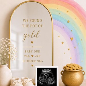 May include: An arched mirror with a gold frame featuring the text WE FOUND THE POT OF gold BABY DUE OCTOBER 2025, accompanied by a white square frame for a sonogram, a small gold pot filled with gold coins, and a pair of white baby booties.