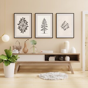 Op de afbeelding: Drie ingelijste kunstdrukken met zwarte frames. De prints tonen een kompasroos, een dennenboom en een bergketen, allemaal in zwart en wit. De prints worden weergegeven boven een houten console.