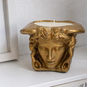 Könnte beinhalten: Ein goldfarbener Kerzenhalter in Form einer klassischen Büste. Der Kerzenhalter hat eine quadratische Basis und ein geformtes Gesicht mit detaillierten Merkmalen. Die Kerze ist weiß, und der Halter steht auf einer weißen Oberfläche.