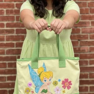 May include: A light yellow tote bag with green accents and handles. The bag features an embroidered image of Tinkerbell with blue wings and floral accents. The bag is held in front of a brick wall.
