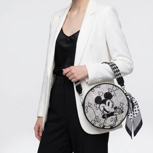 May include: A round, silver glitter crossbody bag featuring a black graphic of Mickey Mouse. The bag has a black and white houndstooth patterned shoulder strap and a black and white polka dot scarf tied to the side attachment ring.
