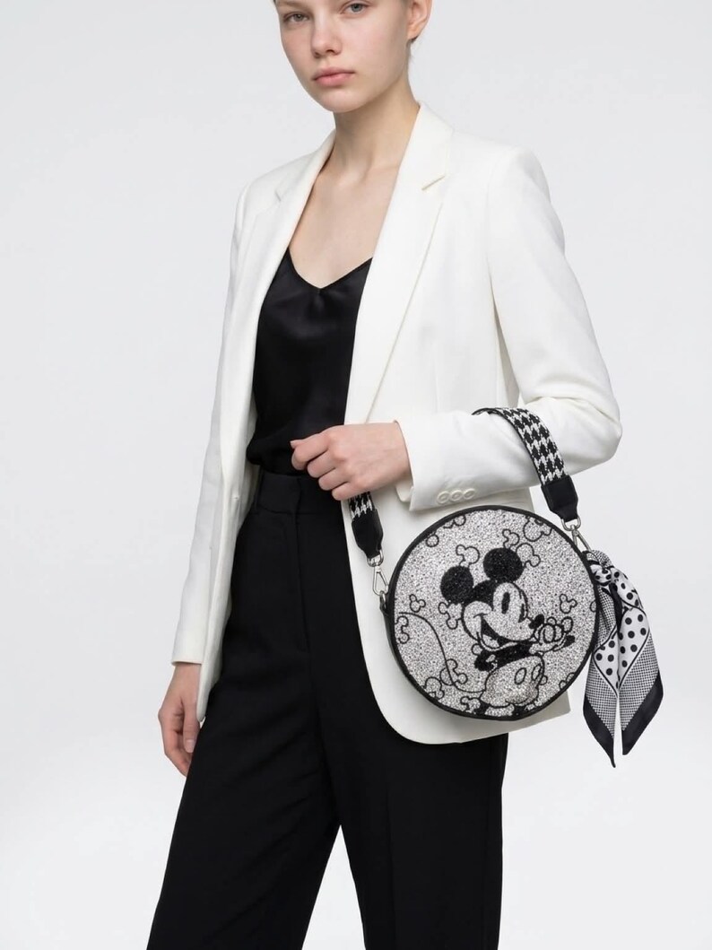May include: A round, silver glitter crossbody bag featuring a black graphic of Mickey Mouse. The bag has a black and white houndstooth patterned shoulder strap and a black and white polka dot scarf tied to the side attachment ring.