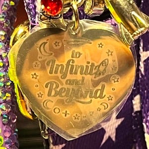Puede incluir: Un dije de acr&iacute;lico transparente en forma de coraz&oacute;n con el texto grabado 'to Infinity and Beyond' rodeado de peque&ntilde;as estrellas y lunas crecientes. El dije est&aacute; unido a una argolla de metal dorado con un brillante rojo y una nave espacial.