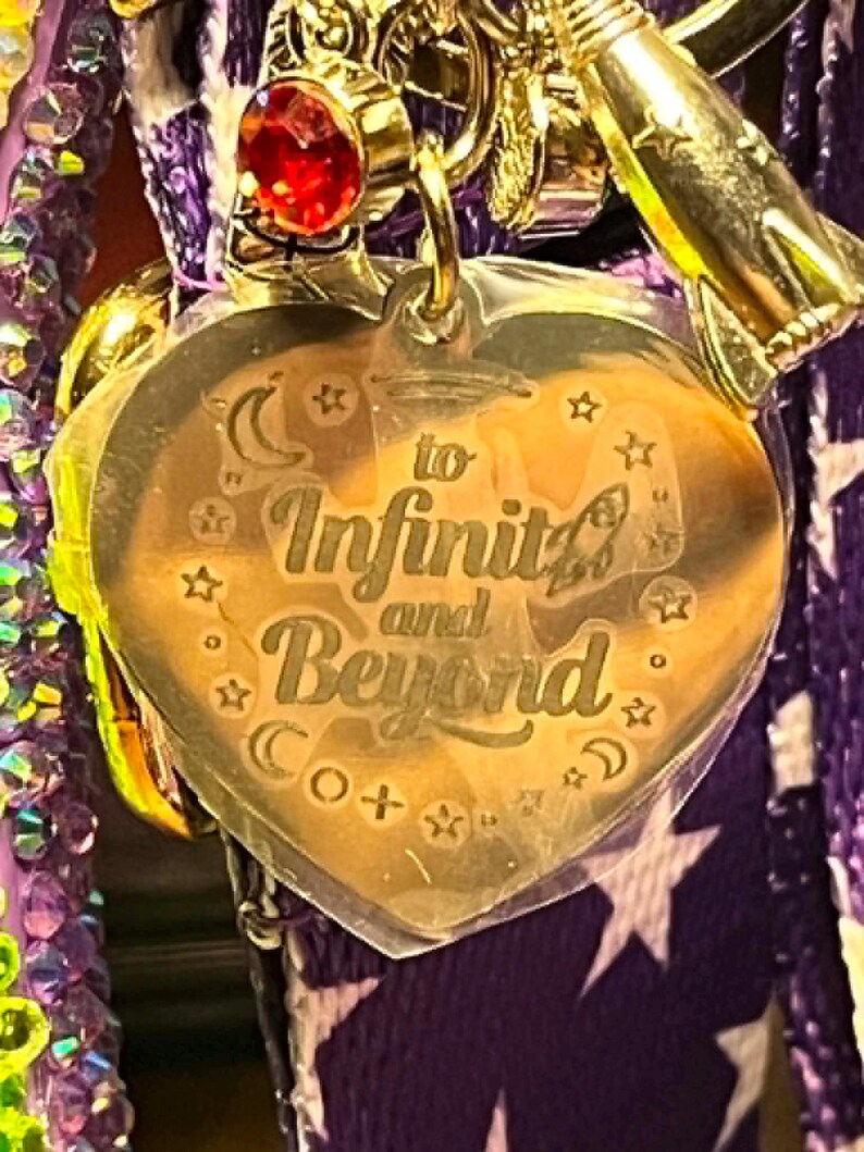 Puede incluir: Un dije de acr&iacute;lico transparente en forma de coraz&oacute;n con el texto grabado 'to Infinity and Beyond' rodeado de peque&ntilde;as estrellas y lunas crecientes. El dije est&aacute; unido a una argolla de metal dorado con un brillante rojo y una nave espacial.