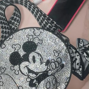 May include: A round crossbody bag featuring a crystal-encrusted Mickey Mouse design in black and clear stones. The bag has a black and white houndstooth patterned strap, a black and white polka dot side bow, a smartphone, and a small lip gloss tube.